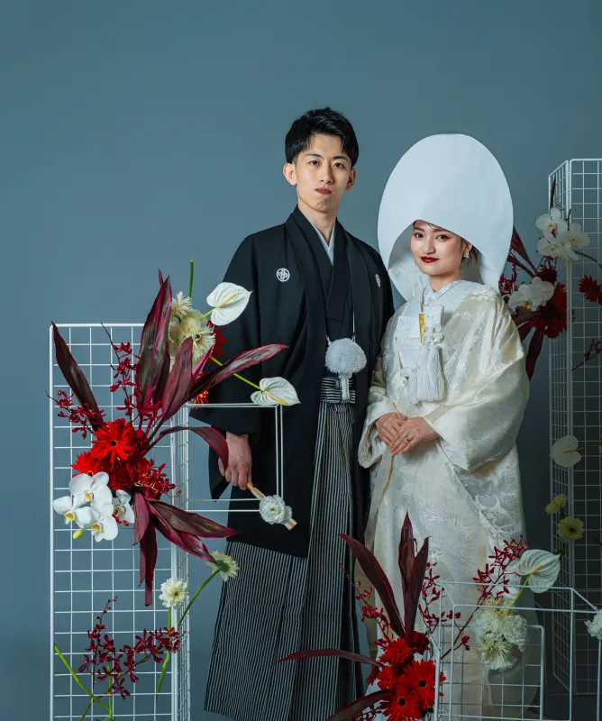 赤い装花を背に並ぶ黒紋付袴の新郎と綿帽子に白無垢の花嫁（和装シーン）