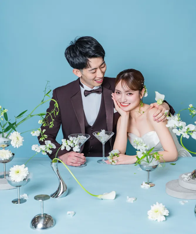 白い装花のテーブルで頬杖をつく花嫁に寄り添う新郎（Table シーン）